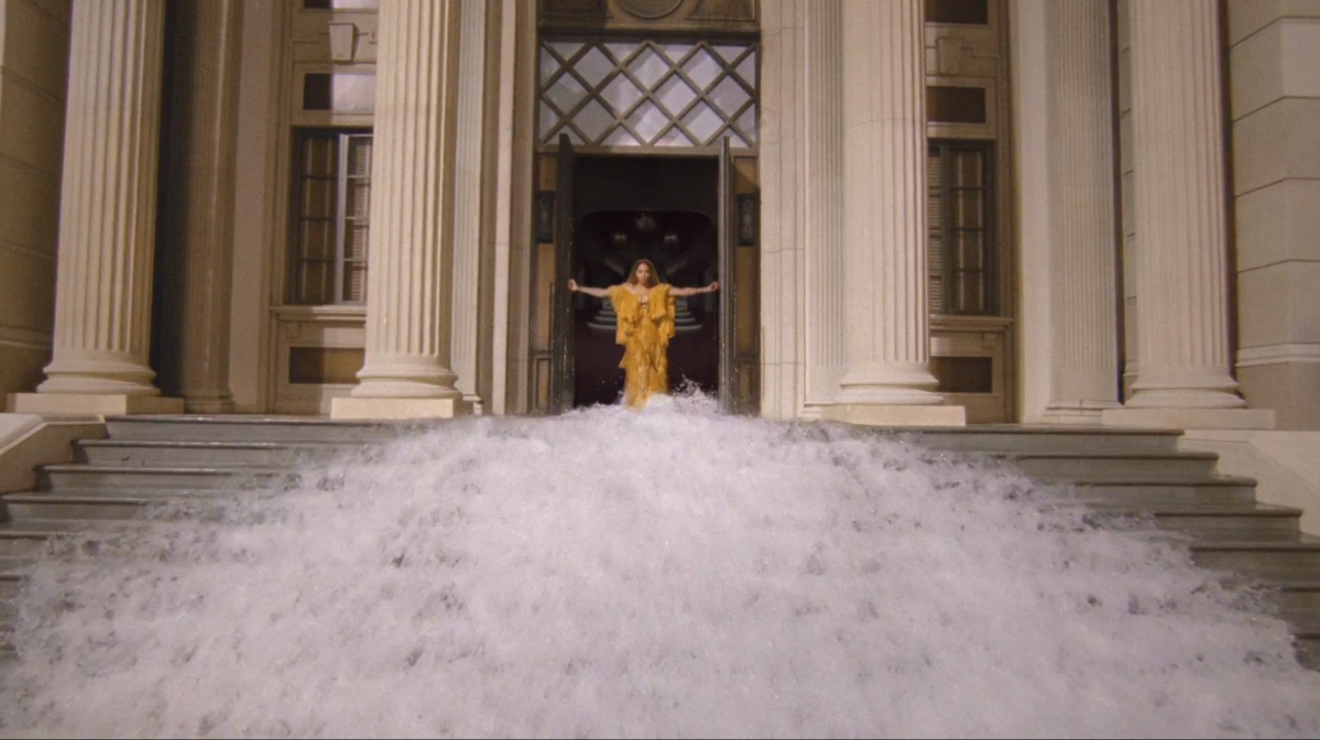Beyonce in a yellow dress stands in an open doorway as water flows down the staircase before her.
