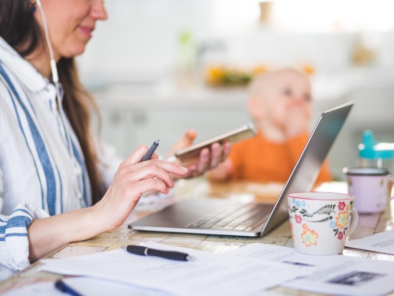 Midsection of working mother using technologies while daughter sitting in background at dining table