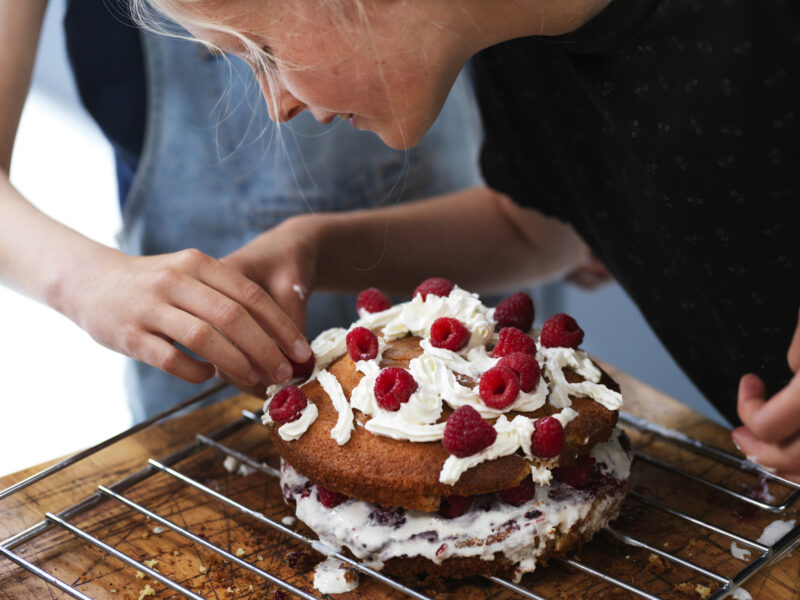 Girl and her sister baking a cake, decorating cake with fresh cream and raspberries at kitchen table, cropped - stock photo