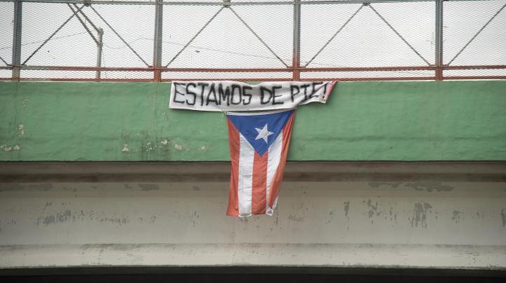 A flag hangs from a highway overpass that reads “Estamos de pie,” in Caguas, Puerto Rico, which is meant to motivate people to stay strong and overcome the disaster left behind Hurricane Maria. Photo by Airman 1st Class Caleb Nunez for  U.S. Air Force. This photo is licensed under CC BY 2.0.