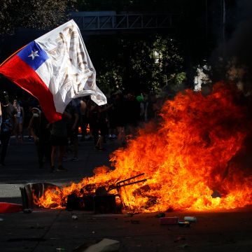 A demonstrator waves a Chilean flag next to a fire as clashing with riot police during a protest against President Sebastian Piñera on October 21, 2019 in Santiago, Chile. Photo by Marcelo Hernandez/Getty Images