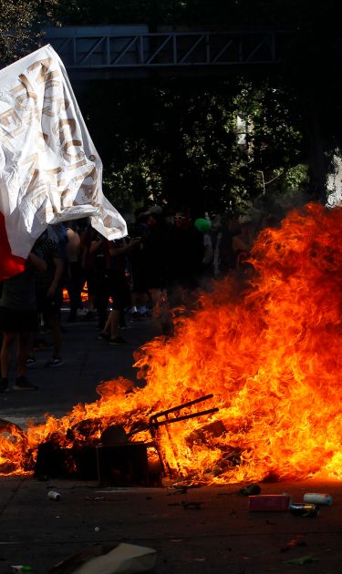 A demonstrator waves a Chilean flag next to a fire as clashing with riot police during a protest against President Sebastian Piñera on October 21, 2019 in Santiago, Chile. Photo by Marcelo Hernandez/Getty Images