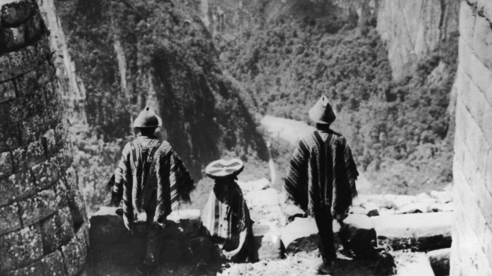 Three Peruvian Indians stand in traditional dress on a masonry ledge of the ancient Incan city of Machu Picchu and look out over a deep valley at one of the Amazon's source streams, early 20th Century. Photo by Henry Clay Gipson/Frederic Lewis/Getty Images