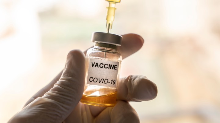 Cropped hand wearing a nitrile glove holding a COVID-19 vaccine vial and a syringe. Getty Images