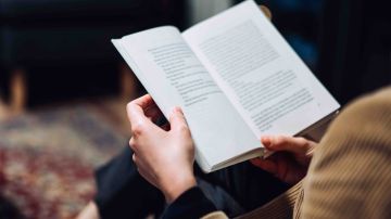 Cropped shot of young Asian woman taking a break from technology, relaxing and reading book on the sofa at a cozy home in the evening