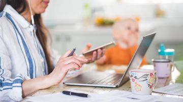 Midsection of working mother using technologies while daughter sitting in background at dining table