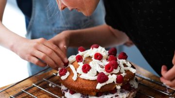 Girl and her sister baking a cake, decorating cake with fresh cream and raspberries at kitchen table, cropped - stock photo