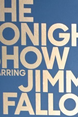 Amaya and Bryan from Love Island USA Season 7 wearing all black and standing next in front of a blue background with the logo for "The Tonight Show starring Jimmy Fallon" in gold lettering
