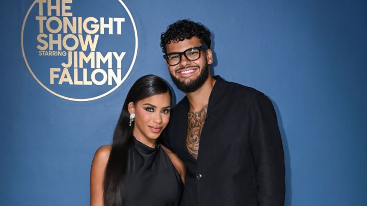 Amaya and Bryan from Love Island USA Season 7 wearing all black and standing next in front of a blue background with the logo for "The Tonight Show starring Jimmy Fallon" in gold lettering