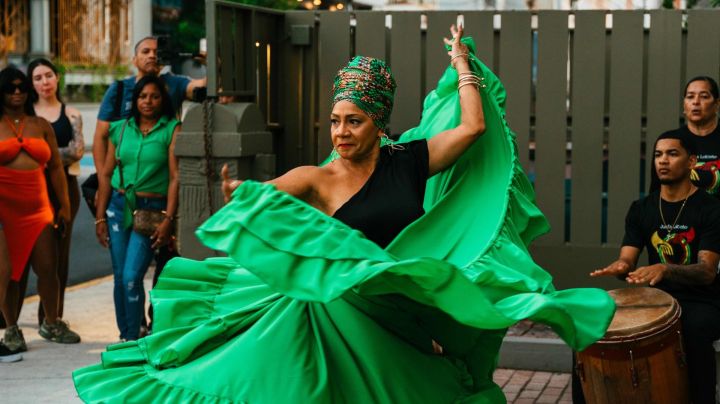 Dancer with a flowing green skirt pictured in the middle with drummers to her right and spectators to her left for La Sala Libre